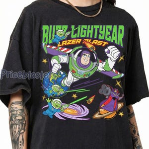 May include: Black t-shirt featuring a colorful graphic of Buzz Lightyear in flight, with the words "Buzz Lightyear Lazer Blast" in green. The design includes space aliens and a villain.