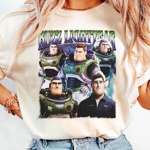 May include: Cream-colored t-shirt featuring a graphic of Buzz Lightyear from Toy Story. The design includes multiple images of Buzz in his spacesuit and the text "BUZZ LIGHTYEAR" in a retro font. The shirt is a casual wear item.