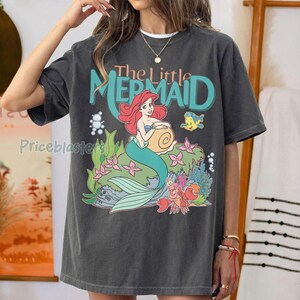 May include: Dark gray t-shirt featuring a colorful graphic of Ariel from The Little Mermaid. The design includes Ariel with red hair and a turquoise tail, along with the text "The Little Mermaid". Additional characters and floral elements are also present.