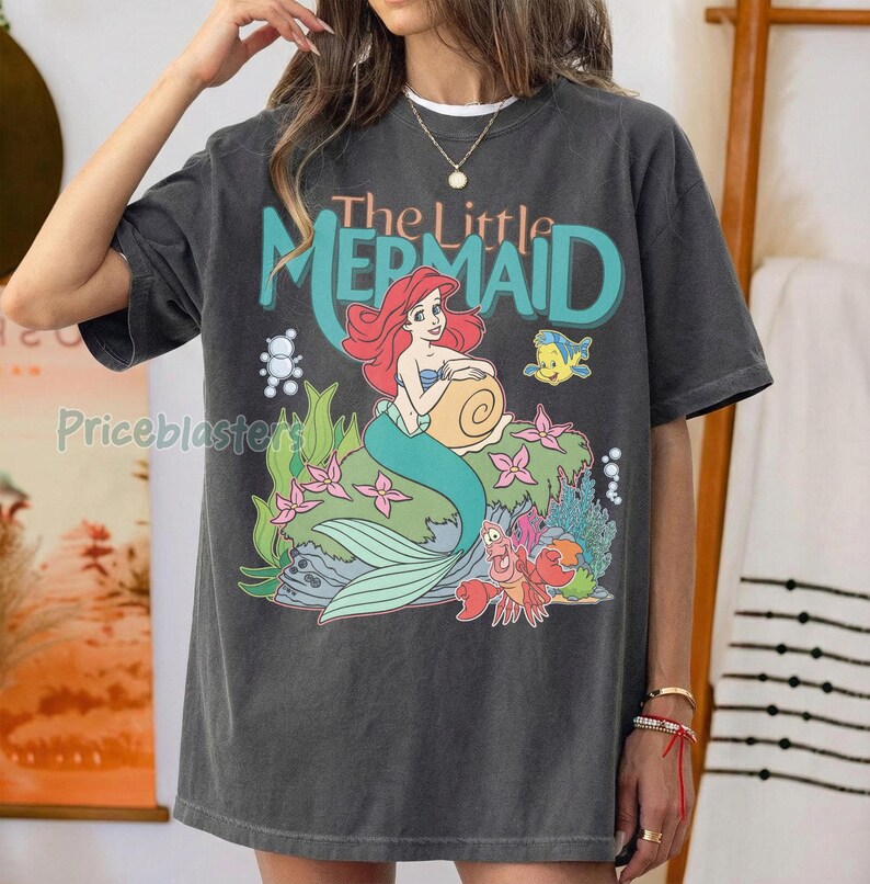 May include: Dark gray t-shirt featuring a colorful graphic of Ariel from The Little Mermaid. The design includes Ariel with red hair and a turquoise tail, along with the text "The Little Mermaid". Additional characters and floral elements are also present.