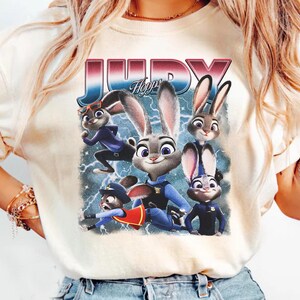 Op de afbeelding: Cr&egrave;mekleurig T-shirt met een afbeelding van het personage Judy Hopps uit de animatiefilm Zootopia. Het ontwerp bevat Judy en andere personages, met de naam "JUDY" in een retro lettertype. De achtergrond heeft bliksem.