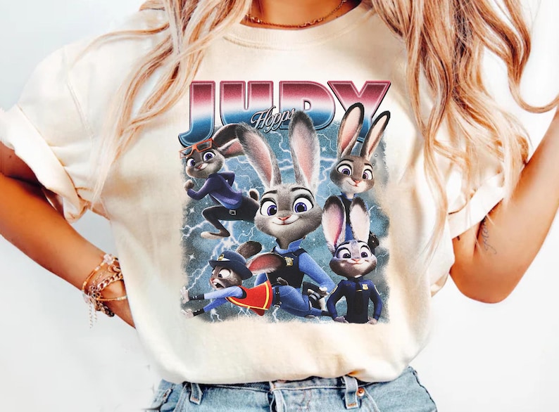 Op de afbeelding: Cr&egrave;mekleurig T-shirt met een afbeelding van het personage Judy Hopps uit de animatiefilm Zootopia. Het ontwerp bevat Judy en andere personages, met de naam "JUDY" in een retro lettertype. De achtergrond heeft bliksem.