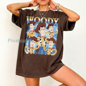 May include: Brown t-shirt featuring a graphic of Woody from Toy Story. The design includes the word "WOODY" in gold lettering above a cartoon image of Woody with a blue lightning background. The shirt has short sleeves.