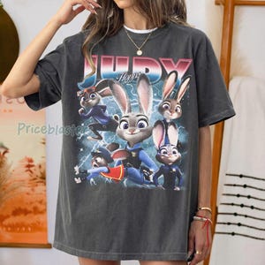 Op de afbeelding: Donkergrijs T-shirt met een afbeelding van Judy Hopps uit Zootopia. Het ontwerp bevat Judy en andere personages met bliksemflitsen en de tekst "JUDY Hopps" in roze en blauw.