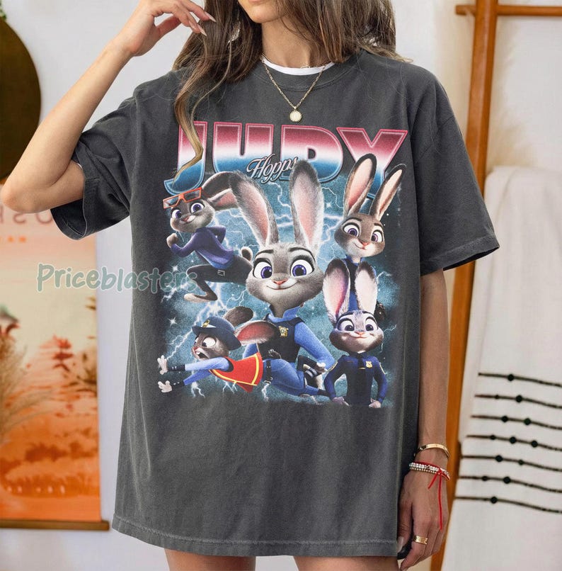 Op de afbeelding: Donkergrijs T-shirt met een afbeelding van Judy Hopps uit Zootopia. Het ontwerp bevat Judy en andere personages met bliksemflitsen en de tekst "JUDY Hopps" in roze en blauw.