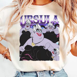 May include: Cream-colored t-shirt featuring a graphic of Ursula from The Little Mermaid. The design includes the word "URSULA" in purple and images of Ursula in various poses, with black and purple accents.