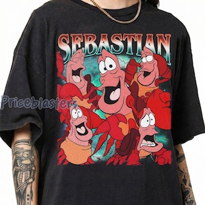 Pu&ograve; includere: T-shirt grigio scuro con una grafica colorata del personaggio Sebastian de La Sirenetta. La parola "SEBASTIAN" &egrave; stampata sopra il personaggio in un font sfumato. La grafica include diversi personaggi di granchi animati.