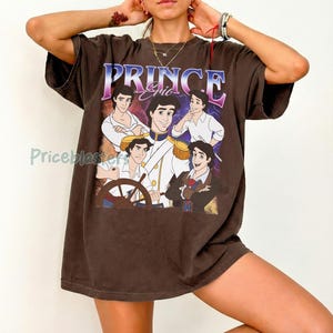 May include: A brown t-shirt featuring a graphic of Prince Eric from The Little Mermaid. The word "PRINCE" is printed in a blue and purple font above the image of the character. The shirt has short sleeves and a relaxed fit.