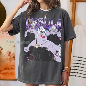 May include: Dark gray t-shirt featuring a graphic of Ursula from The Little Mermaid. The design includes the word "URSULA" in a purple gradient font above the character. The character is depicted in a purple and black color scheme.