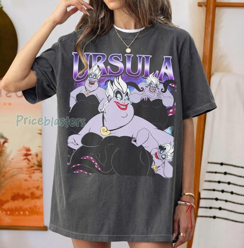 May include: Dark gray t-shirt featuring a graphic of Ursula from The Little Mermaid. The design includes the word "URSULA" in a purple gradient font above the character. The character is depicted in a purple and black color scheme.