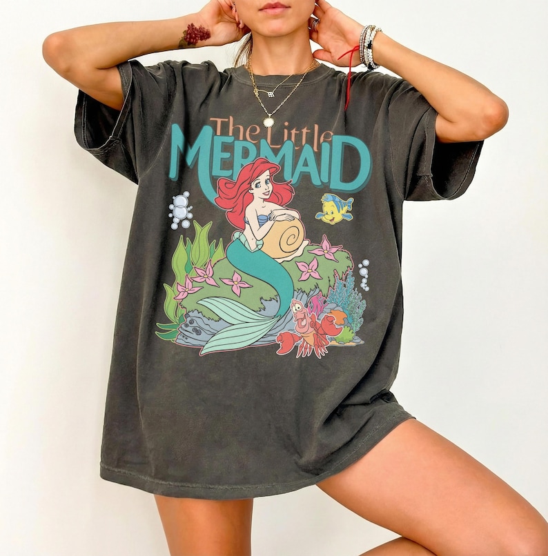 May include: Dark gray t-shirt featuring a graphic of Ariel from The Little Mermaid. The design includes Ariel, Flounder, and Sebastian, with the words "The Little Mermaid" in teal and red. The shirt has a vintage, worn look.