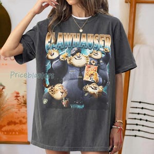 Puede incluir: Camiseta gris oscuro con un dise&ntilde;o de Clawhauser de Zootr&oacute;polis. El dise&ntilde;o incluye a Clawhauser con uniforme de polic&iacute;a, sosteniendo una bolsa de Lucky Chom, y la palabra "Clawhauser". La camiseta tiene un aspecto vintage.