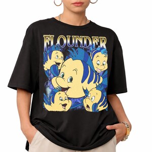 May include: Black t-shirt featuring a graphic of the character Flounder from The Little Mermaid. The design includes multiple images of Flounder in yellow and blue, with the name "FLOUNDER" in gold lettering above the character.