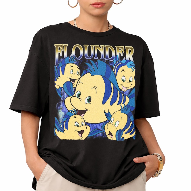 May include: Black t-shirt featuring a graphic of the character Flounder from The Little Mermaid. The design includes multiple images of Flounder in yellow and blue, with the name "FLOUNDER" in gold lettering above the character.