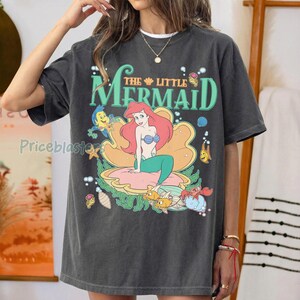 May include: Dark grey t-shirt with a colourful graphic of Ariel from Disney's The Little Mermaid. The design features Ariel on a seashell, surrounded by sea creatures and the film title in green.
