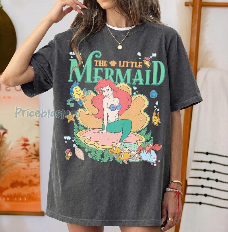 May include: Dark grey t-shirt with a colourful graphic of Ariel from Disney's The Little Mermaid. The design features Ariel on a seashell, surrounded by sea creatures and the film title in green.