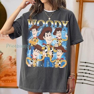 May include: Dark gray t-shirt featuring a graphic of the character Woody from Toy Story. The design includes multiple Woody figures, the word "WOODY" in gold lettering, and a blue lightning bolt background. The shirt is a relaxed fit.