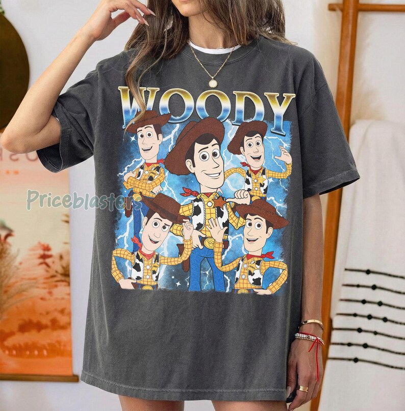 May include: Dark gray t-shirt featuring a graphic of the character Woody from Toy Story. The design includes multiple Woody figures, the word "WOODY" in gold lettering, and a blue lightning bolt background. The shirt is a relaxed fit.