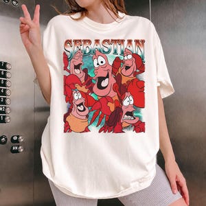 Pu&ograve; includere: T-shirt color crema con una grafica del personaggio Sebastian de La Sirenetta. Il design include la parola "Sebastian" in un font retr&ograve;, con diversi granchi dei cartoni animati in varie pose. La maglietta &egrave; di vestibilit&agrave; ampia.