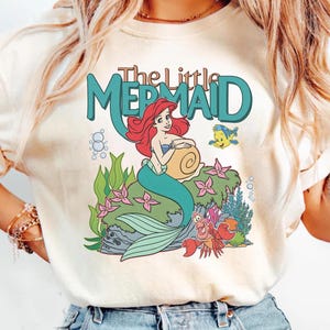 May include: Cream-colored t-shirt featuring a colorful graphic of Ariel from The Little Mermaid. The design includes Ariel with red hair, a teal tail, and the text "The Little Mermaid" in teal. Other characters include Flounder and Sebastian.