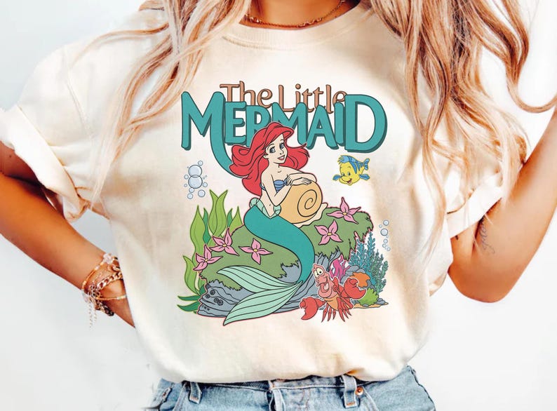 May include: Cream-colored t-shirt featuring a colorful graphic of Ariel from The Little Mermaid. The design includes Ariel with red hair, a teal tail, and the text "The Little Mermaid" in teal. Other characters include Flounder and Sebastian.