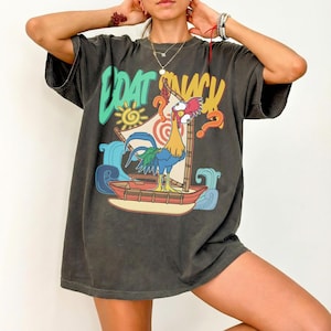 Puede incluir: Camiseta gris oscuro con un gr&aacute;fico colorido de un gallo navegando en un barco, con las palabras "BOAT SNACK" en una fuente l&uacute;dica. El dise&ntilde;o incluye olas, un sol y signos de interrogaci&oacute;n.