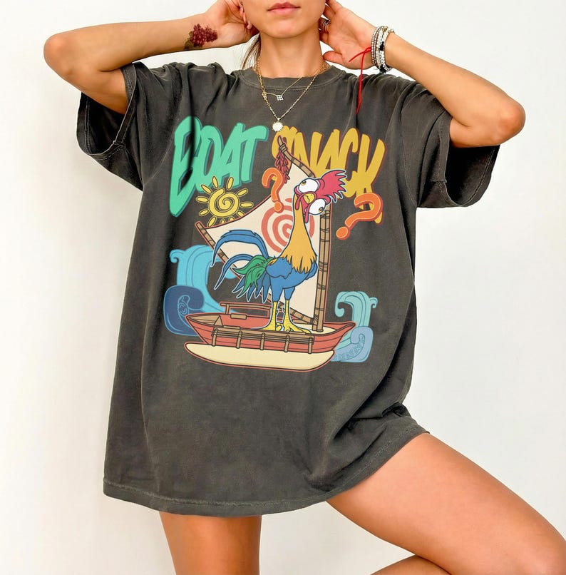 Puede incluir: Camiseta gris oscuro con un gr&aacute;fico colorido de un gallo navegando en un barco, con las palabras "BOAT SNACK" en una fuente l&uacute;dica. El dise&ntilde;o incluye olas, un sol y signos de interrogaci&oacute;n.