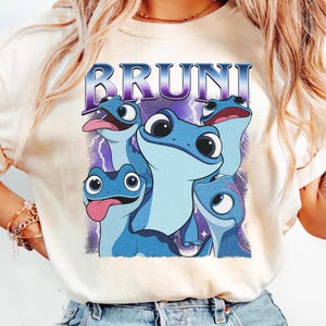 May include: Cream-colored t-shirt featuring a graphic of five blue salamanders with large eyes and tongues sticking out. The word "BRUNI" is printed above the salamanders in a purple and blue font. The design is set against a purple lightning background.