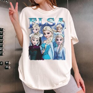 May include: Cream-colored t-shirt featuring the word "ELSA" in blue, with images of the character Elsa from the movie Frozen. The graphic includes multiple depictions of Elsa in various poses and outfits.