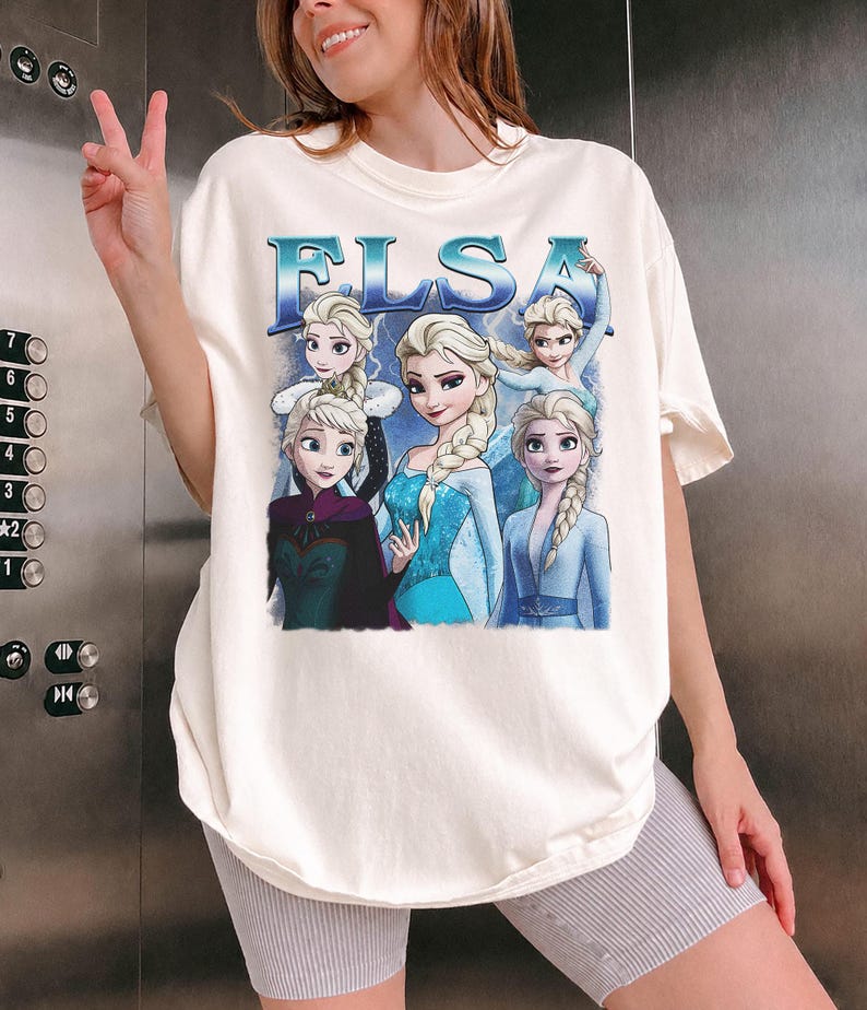 May include: Cream-colored t-shirt featuring the word "ELSA" in blue, with images of the character Elsa from the movie Frozen. The graphic includes multiple depictions of Elsa in various poses and outfits.
