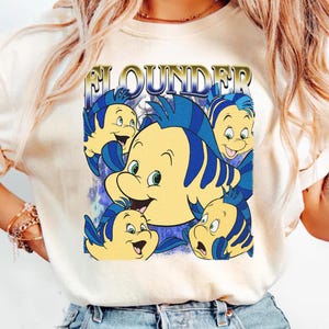 May include: Cream-colored t-shirt featuring a graphic of the character Flounder from The Little Mermaid. The design includes multiple yellow and blue fish with the word "Flounder" in gold lettering. The shirt is a casual style.