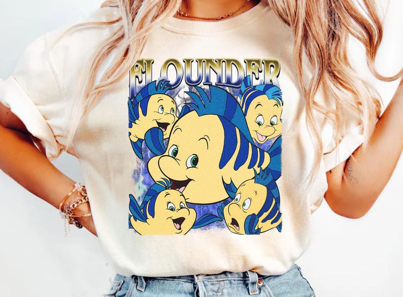 May include: Cream-colored t-shirt featuring a graphic of the character Flounder from The Little Mermaid. The design includes multiple yellow and blue fish with the word "Flounder" in gold lettering. The shirt is a casual style.