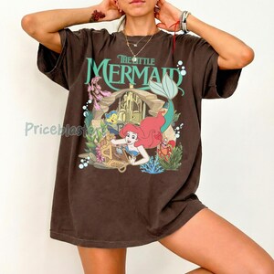 Może przedstawiać: Brązowa koszulka z grafiką z Małej Syrenki Disneya. Projekt zawiera Ariel, Florka, Sebastiana i napis "The Little Mermaid" w kolorze turkusowym. Koszulka ma kr&oacute;tkie rękawy i luźny kr&oacute;j.