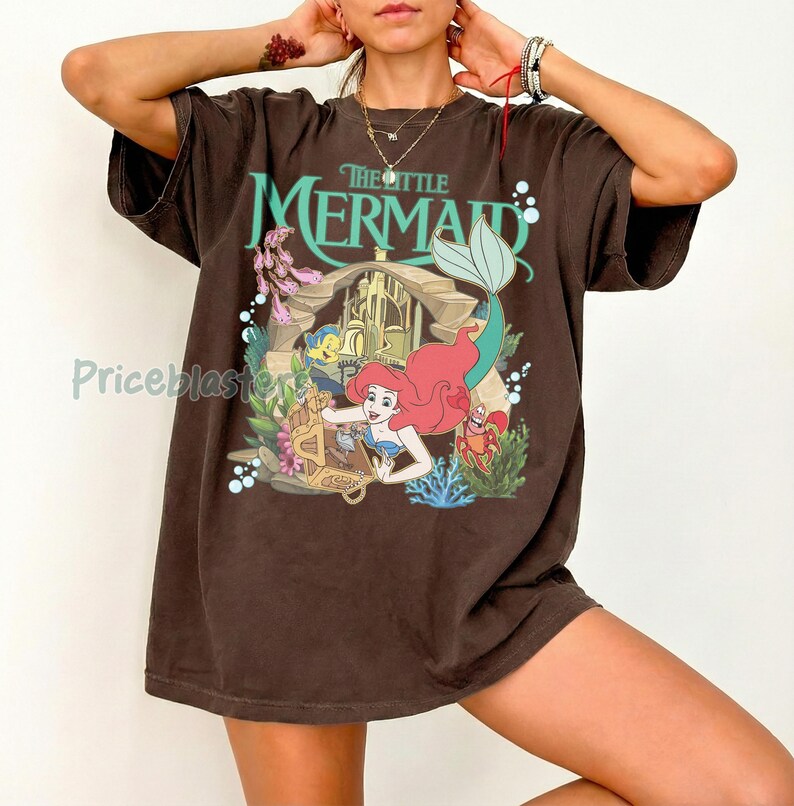 Może przedstawiać: Brązowa koszulka z grafiką z Małej Syrenki Disneya. Projekt zawiera Ariel, Florka, Sebastiana i napis "The Little Mermaid" w kolorze turkusowym. Koszulka ma kr&oacute;tkie rękawy i luźny kr&oacute;j.