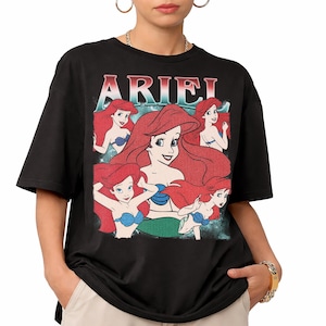 Pu&ograve; includere: T-shirt nera con grafica di Ariel de La Sirenetta. Il design include pi&ugrave; immagini di Ariel con capelli rossi e reggiseno a conchiglia blu, con la scritta "ARIEL" in testo rosso e bianco nella parte superiore.