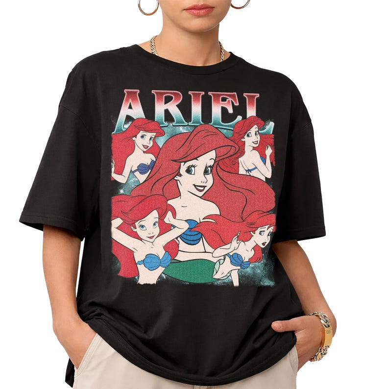 Pu&ograve; includere: T-shirt nera con grafica di Ariel de La Sirenetta. Il design include pi&ugrave; immagini di Ariel con capelli rossi e reggiseno a conchiglia blu, con la scritta "ARIEL" in testo rosso e bianco nella parte superiore.