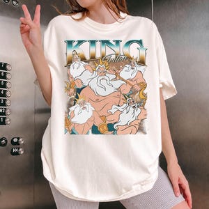 May include: Cream-colored t-shirt featuring a graphic of King Triton from The Little Mermaid. The design includes the text "KING Triton" in a retro font, with illustrations of the character in various poses. The shirt has short sleeves and a relaxed fit.