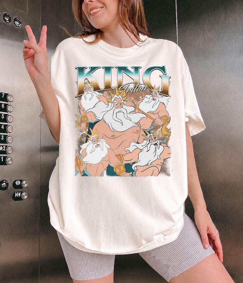 May include: Cream-colored t-shirt featuring a graphic of King Triton from The Little Mermaid. The design includes the text "KING Triton" in a retro font, with illustrations of the character in various poses. The shirt has short sleeves and a relaxed fit.