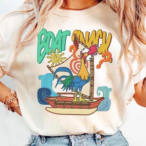 Puede incluir: Camiseta color crema con un gallo de dibujos animados en un barco con las palabras "BOAT SNACK". El dise&ntilde;o incluye un sol, olas y signos de interrogaci&oacute;n, creando un ambiente tropical divertido. El gallo es colorido, con plumas azules, amarillas y rojas.