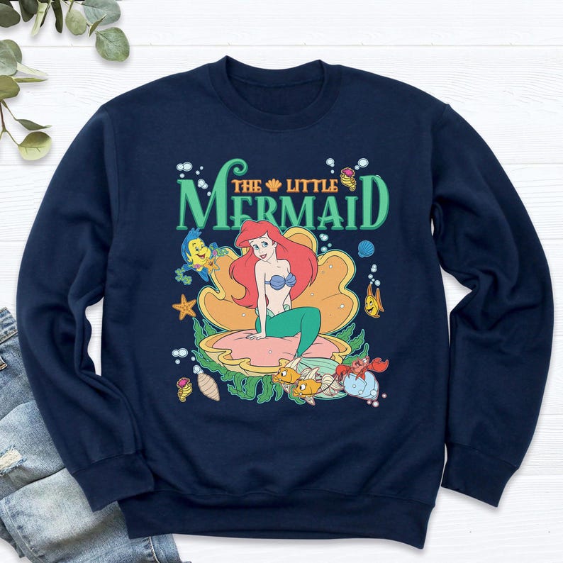 May include: Navy blue sweatshirt with a graphic of Ariel from The Little Mermaid. Ariel, with red hair and a green tail, sits in a seashell. The design includes sea creatures and the words "The Little Mermaid" in green.