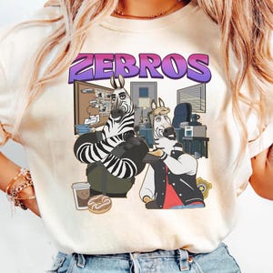 Pu&ograve; includere: T-shirt color crema con grafica a cartoni animati di due zebre in un ambiente d'ufficio. La parola "ZEBROS" &egrave; scritta in viola e blu sopra le zebre. Una zebra indossa una giacca, l'altra un abito a righe bianche e nere.