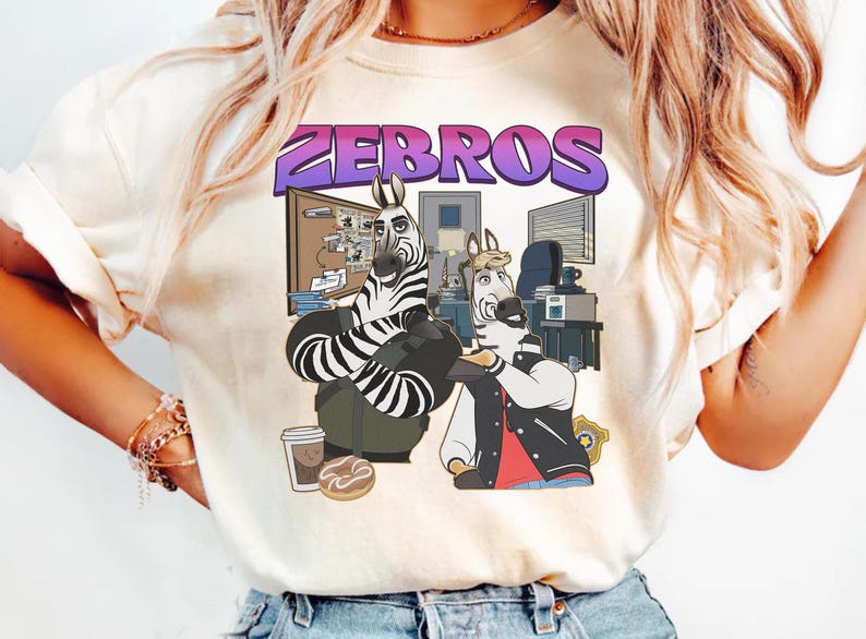 Pu&ograve; includere: T-shirt color crema con grafica a cartoni animati di due zebre in un ambiente d'ufficio. La parola "ZEBROS" &egrave; scritta in viola e blu sopra le zebre. Una zebra indossa una giacca, l'altra un abito a righe bianche e nere.