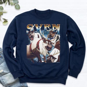 May include: Navy blue sweatshirt featuring a graphic of Sven from the movie Frozen. The graphic includes multiple reindeer with the word "SVEN" in a gold and blue font. The sweatshirt is made of a soft material.