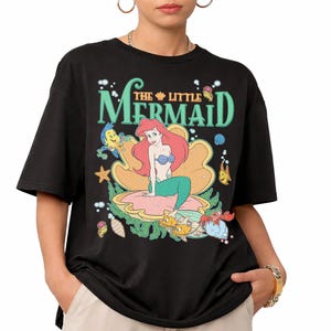 May include: Black t-shirt with a colourful graphic of Ariel from Disney's The Little Mermaid. The design shows Ariel in a seashell, surrounded by sea creatures, with the film title in green and teal lettering.