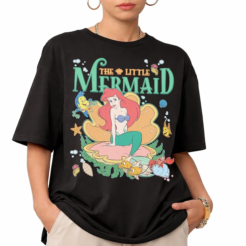 May include: Black t-shirt with a colourful graphic of Ariel from Disney's The Little Mermaid. The design shows Ariel in a seashell, surrounded by sea creatures, with the film title in green and teal lettering.