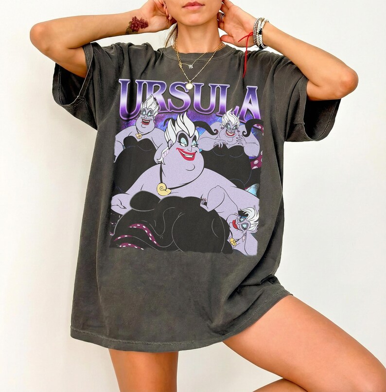 May include: Dark gray t-shirt featuring a graphic of Ursula from The Little Mermaid, with the name "URSULA" in purple letters. The design includes Ursula's face and tentacles. The shirt has short sleeves and a relaxed fit.