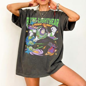May include: Dark gray t-shirt featuring a graphic of Buzz Lightyear with the text "Buzz Lightyear Lazer Blast." The design includes cartoon characters and space-themed elements in green, purple, and blue. The shirt has short sleeves and a relaxed fit.