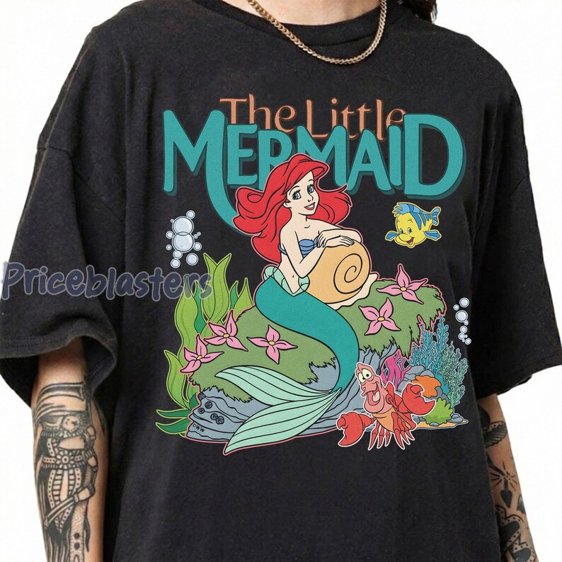 May include: Black t-shirt featuring a colorful graphic of Ariel from The Little Mermaid. The design includes Ariel with red hair, a teal tail, and the text "The Little Mermaid". Other characters include Flounder and Sebastian.