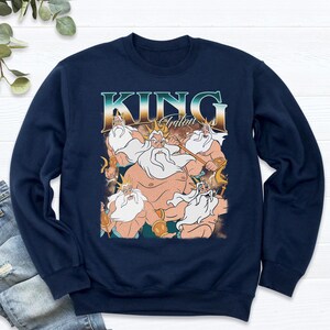 May include: Navy blue sweatshirt featuring a graphic of King Triton from The Little Mermaid. The design includes the text "KING Triton" in a vintage style font, with multiple images of the character. The sweatshirt is a crew neck style.