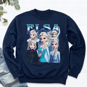 May include: Navy blue sweatshirt featuring a graphic of Elsa from the movie Frozen. The graphic includes multiple images of Elsa with the word "ELSA" in a light blue font. The sweatshirt is made of a soft material.